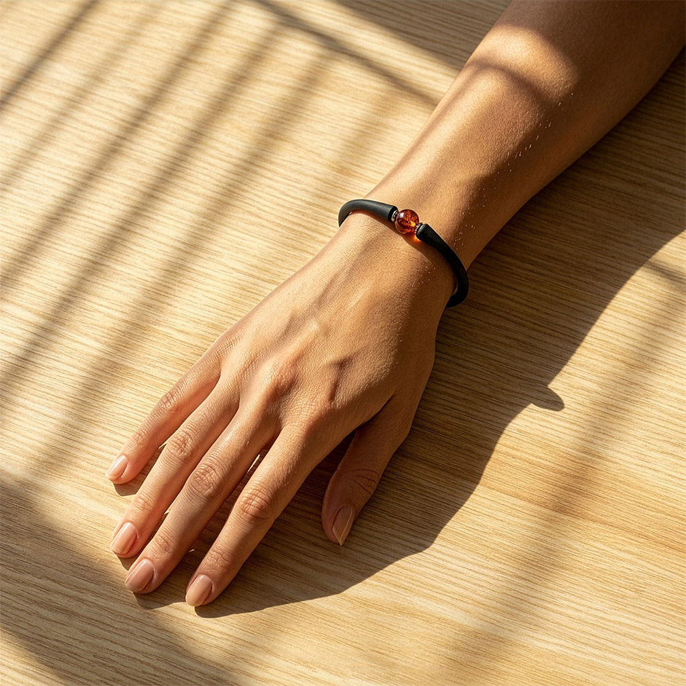 Hand mit schwarzem Armband und Bernstein-Perle auf heller Holzoberfläche im warmen Sonnenlicht mit weichen Schattenlinien.