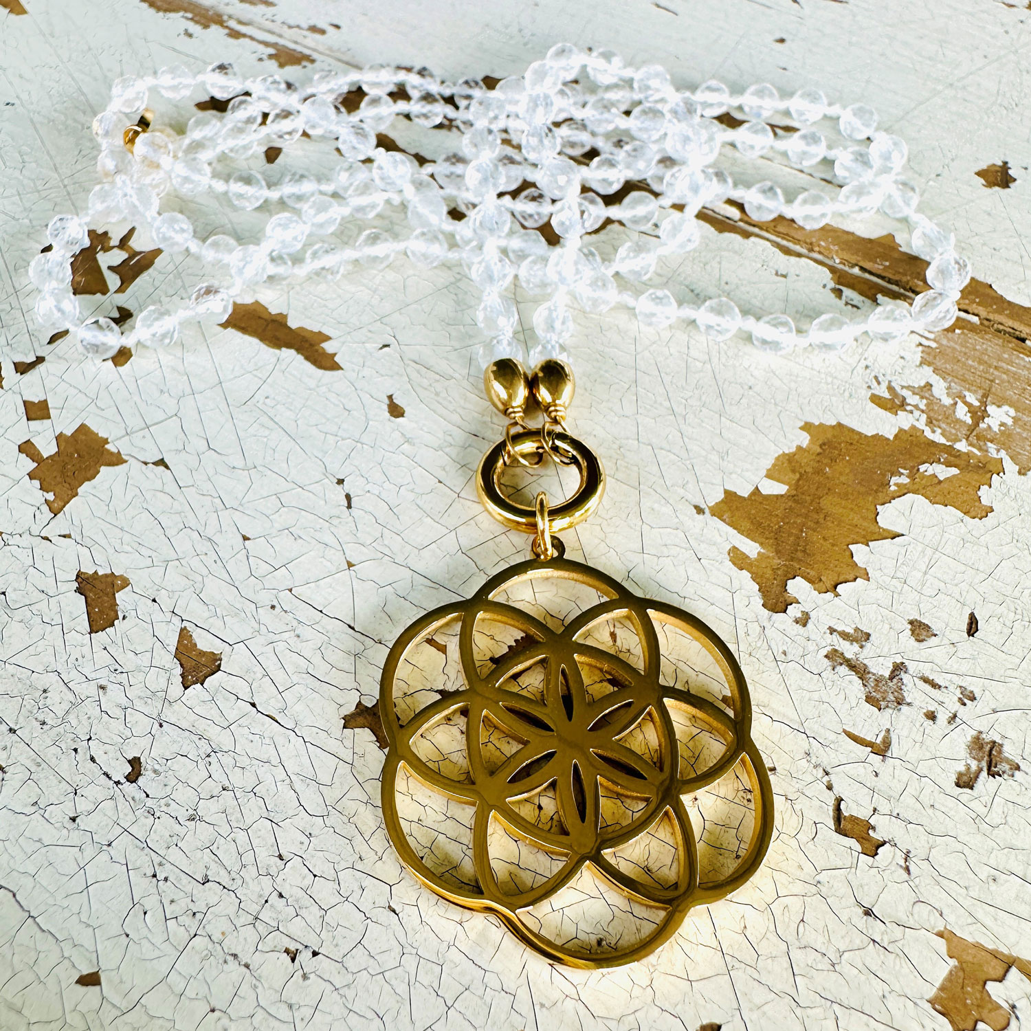 Goldenes, florales Medaillon mit geometrischem Muster an einer Kette aus transparenten Perlen, liegend auf abgeblättertem, weißem Holz.