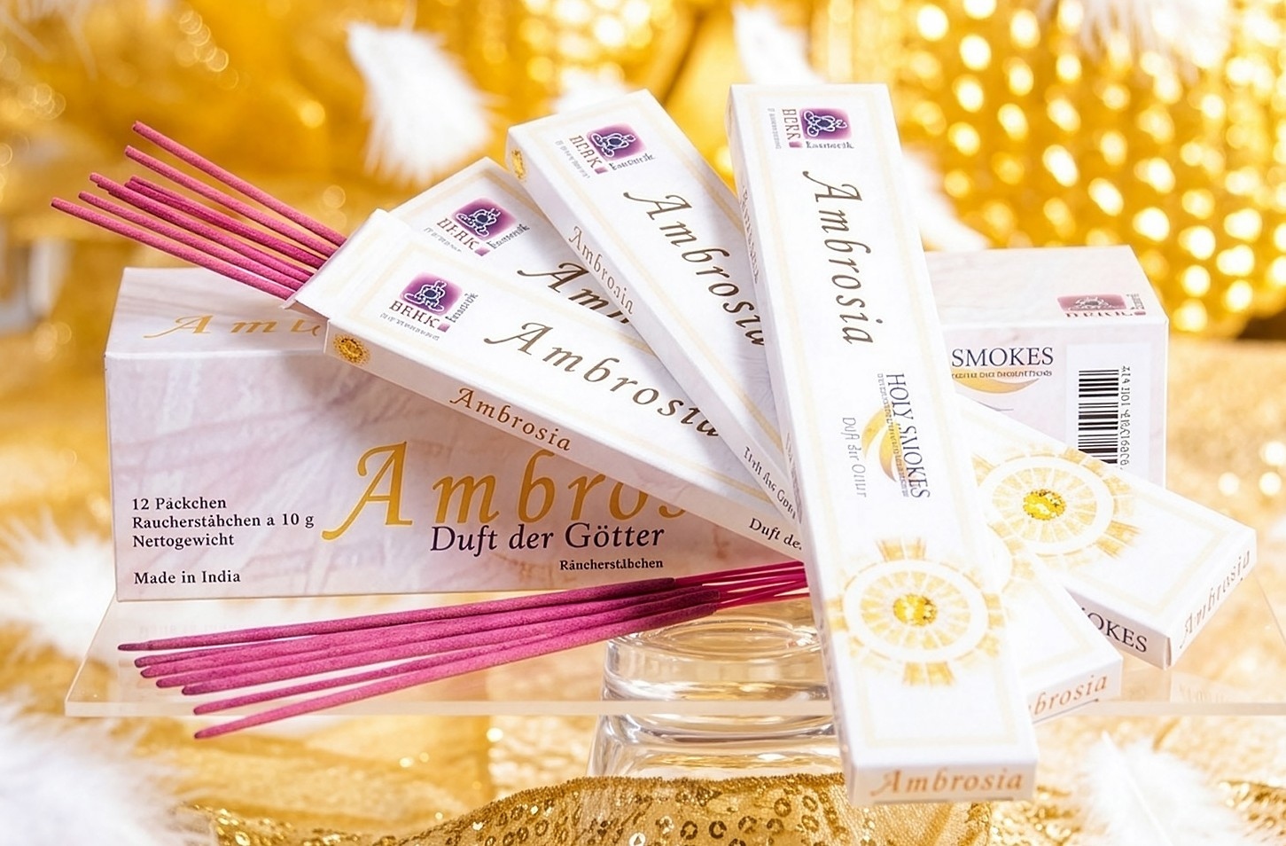 Mehrere Packungen und pinkfarbene Räucherstäbchen der Sorte „Ambrosia – Duft der Götter“ sind dekorativ auf einer goldfarbenen Unterlage angeordnet.