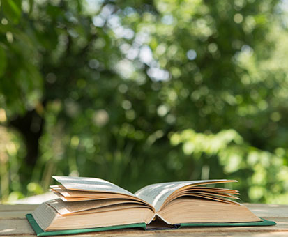 Aufgeschlagenes Buch auf einem Holztisch im Grünen, unscharfer Hintergrund mit Bäumen und Sonnenlicht.