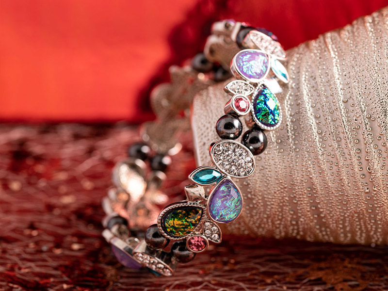Ein elegantes Armband mit tropfenförmigen, schimmernden Schmucksteinen in verschiedenen Farben. Die Steine sind in einer filigranen Silberfassung eingefasst und mit kleinen schwarzen Perlen kombiniert.