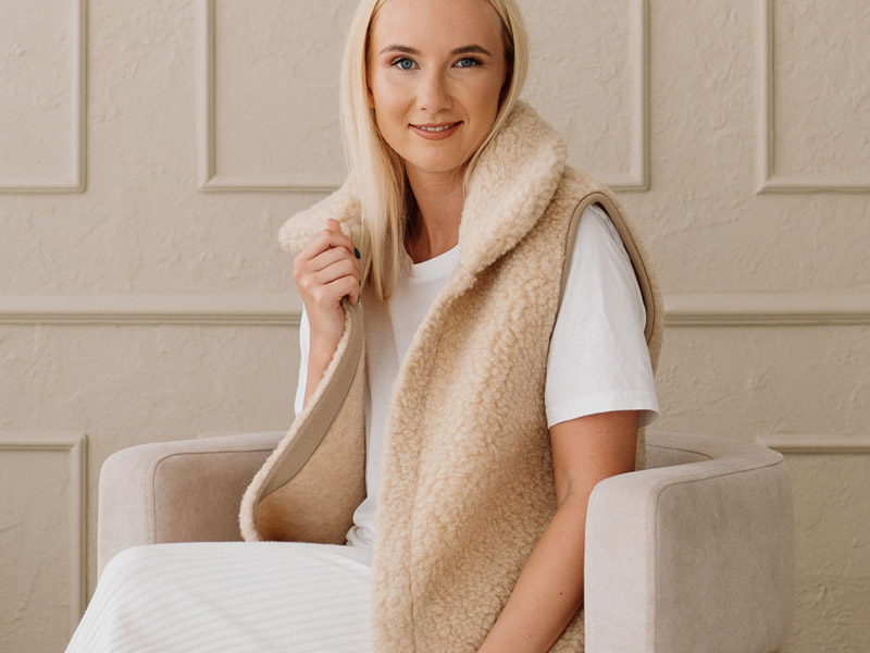 Eine junge Frau sitzt entspannt in einem beigen Sessel und trägt eine warme, naturfarbene Weste aus flauschiger Merinowolle über einem weißen Outfit. 
