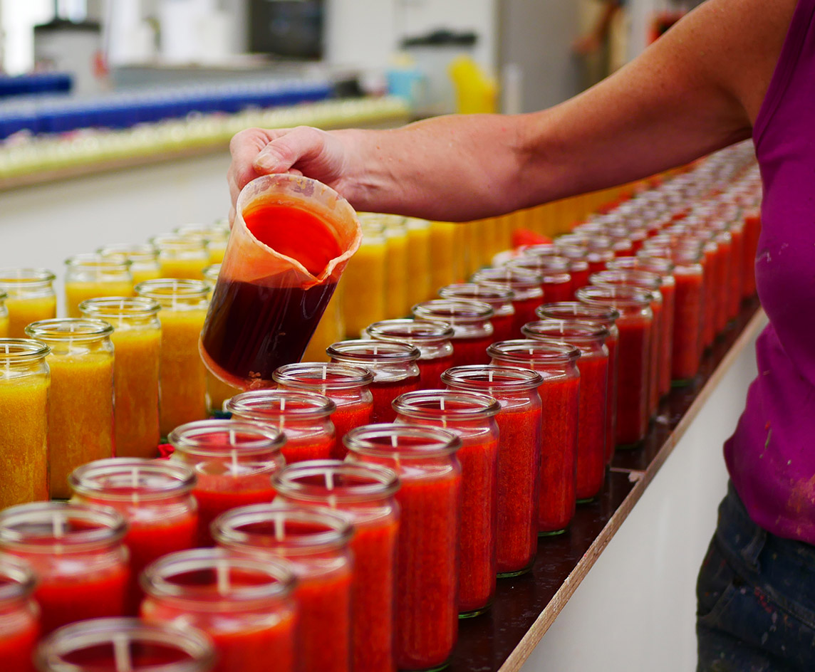 Person gießt farbiges Wachs in Glasbehälter – Herstellung bunter Kerzen in einer Kerzenmanufaktur.