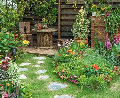 Blühender Garten mit runder Holzsitzgruppe auf kleiner Terrasse, umgeben von Blumenbeeten und Trittsteinen auf dem Rasen.
