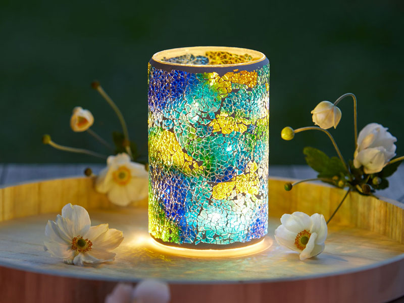 Ein leuchtendes LED-Windlicht steht auf einem Tablett, umgeben von zarten Blüten. Das Windlicht besteht aus einem zylindrischen Mosaikglas mit bunten, rissartig angeordneten Fragmenten in Blau, Grün, Gelb und Türkis.