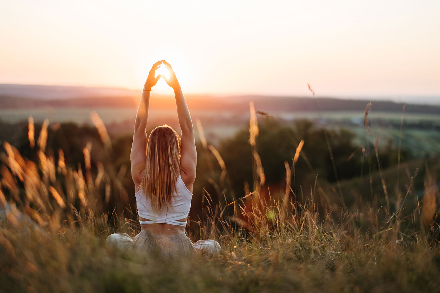 Frau sitzt im hohen Gras auf einem Hügel bei Sonnenuntergang und streckt die Arme zur Sonne empor, mit weiter Landschaft im warmen Abendlicht im Hintergrund.