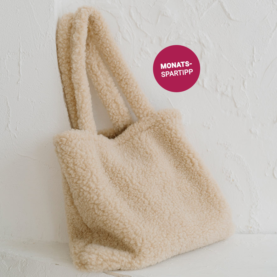 Eine flauschige, beigefarbene Tasche aus weicher Merinowolle lehnt lässig an einer weiß verputzten Wand.