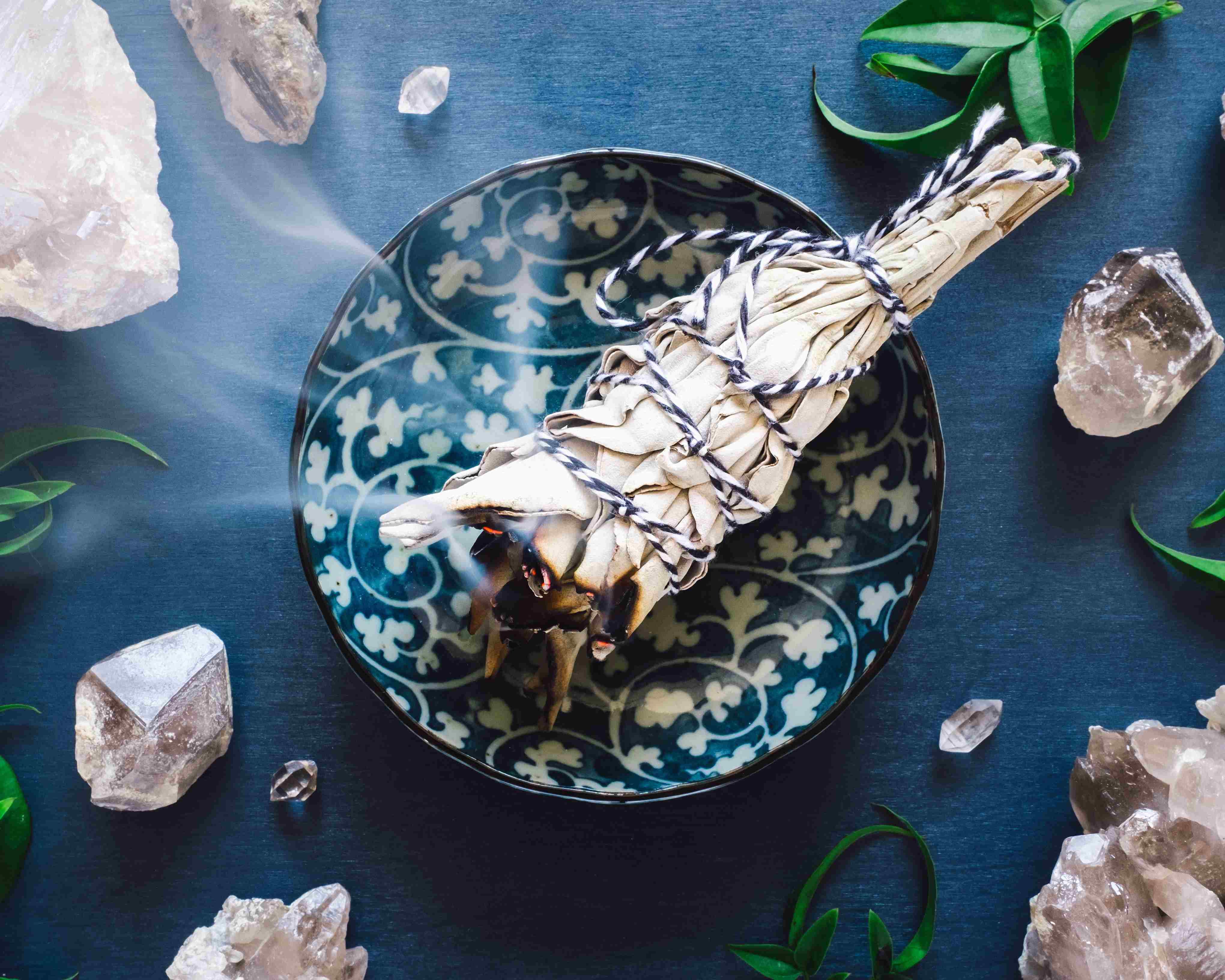 Räucherbündel mit aufsteigendem Rauch liegt in einer gemusterten Schale, umgeben von Kristallen und grünen Blättern auf dunklem Untergrund.