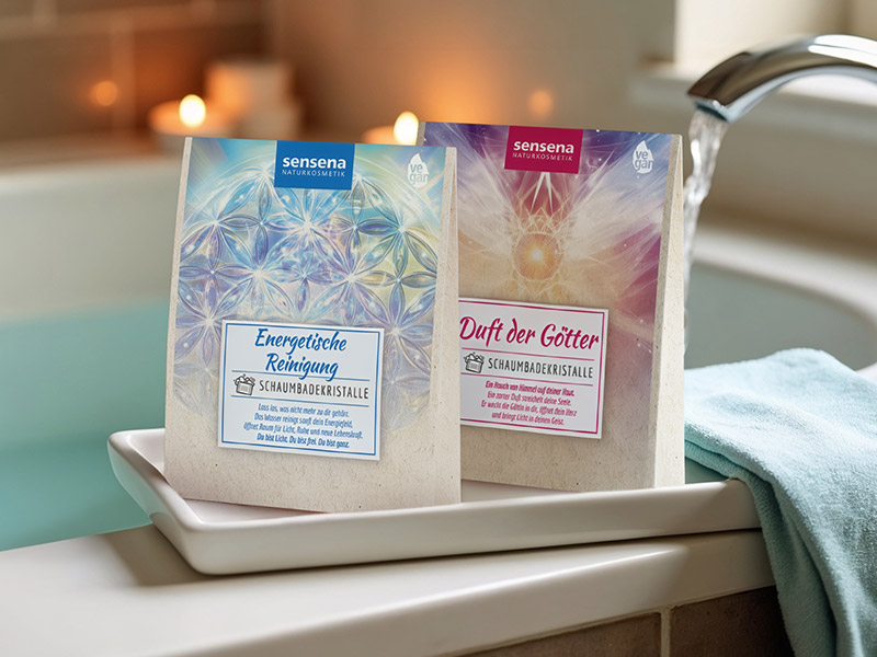 Zwei Packungen Schaumbadekristalle von Sensena mit den Sorten „Energetische Reinigung“ und „Duft der Götter“ stehen am Rand einer gefüllten Badewanne, im Hintergrund Kerzenlicht und fließendes Wasser sorgen für eine entspannte Atmosphäre.