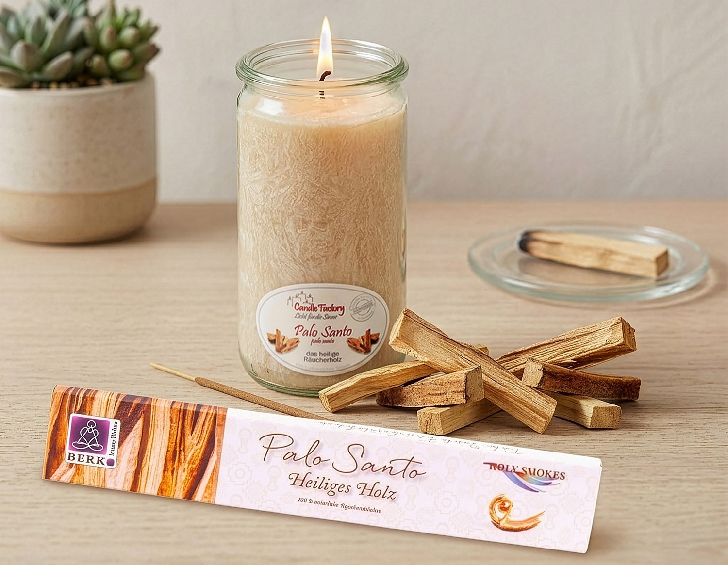 Eine brennende Palo-Santo-Duftkerze steht neben Holzstückchen und einer Packung „Palo Santo Heiliges Holz“ auf einem hellen Tisch.
