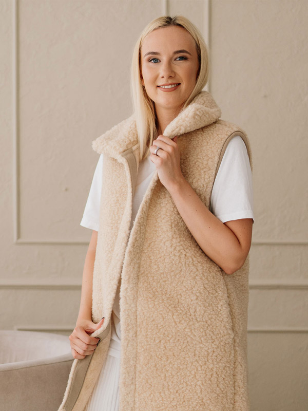 Eine junge Frau sitzt entspannt in einem beigen Sessel und trägt eine warme, naturfarbene Weste aus flauschiger Merinowolle über einem weißen Outfit. 