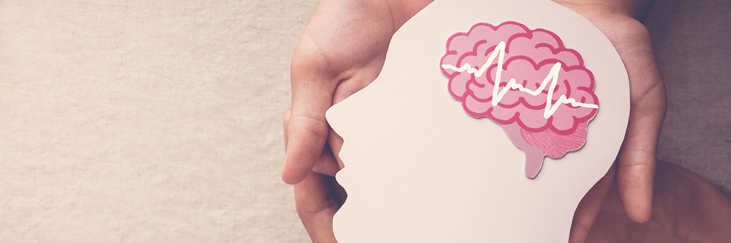 Hände halten eine aus beiger Pappe ausgeschnittene Silhouette eines Kopfes. Darauf liegt eine aus rosa Papier ausgeschnittene stilisierte Darstellung eines Gehirns.