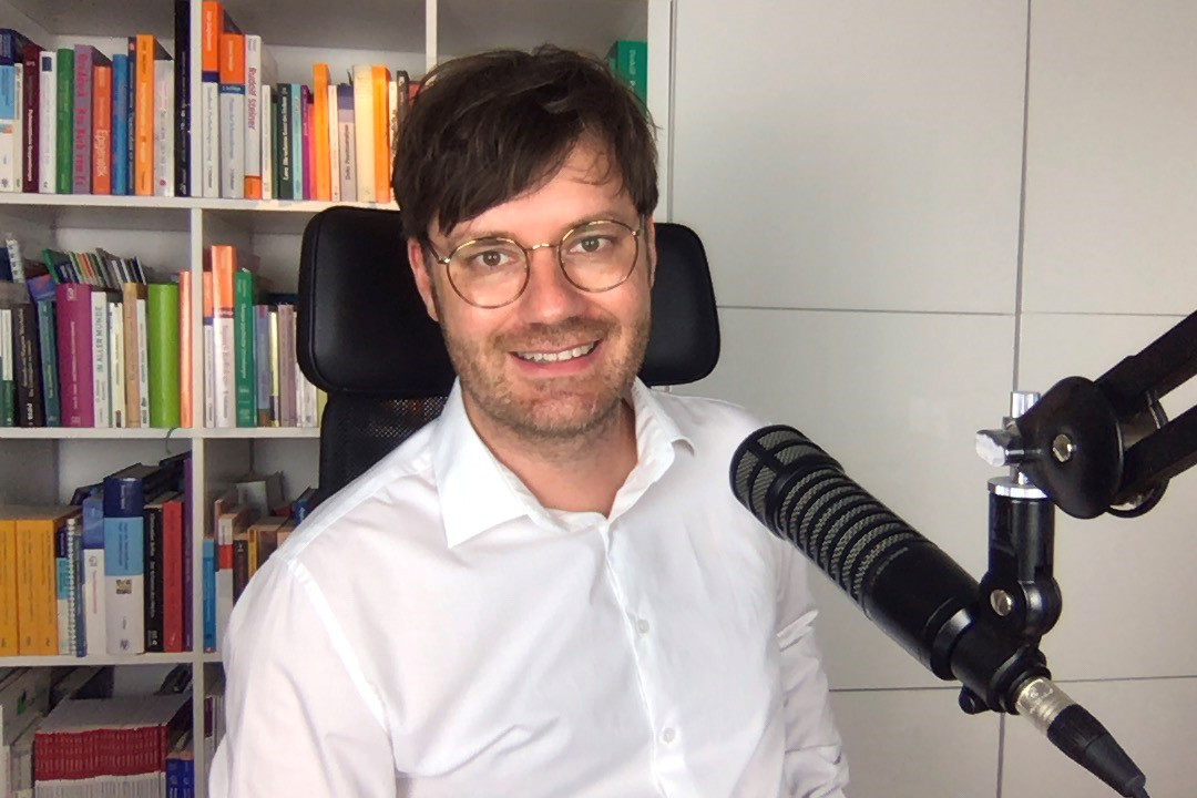 Dr. med. Alexander Kugelstadt trägt ein weißes Hemd und sitzt vor einem Mikrofon. Er lächelt in die Kamera. Im Hintergrund steht ein Bücherregal.