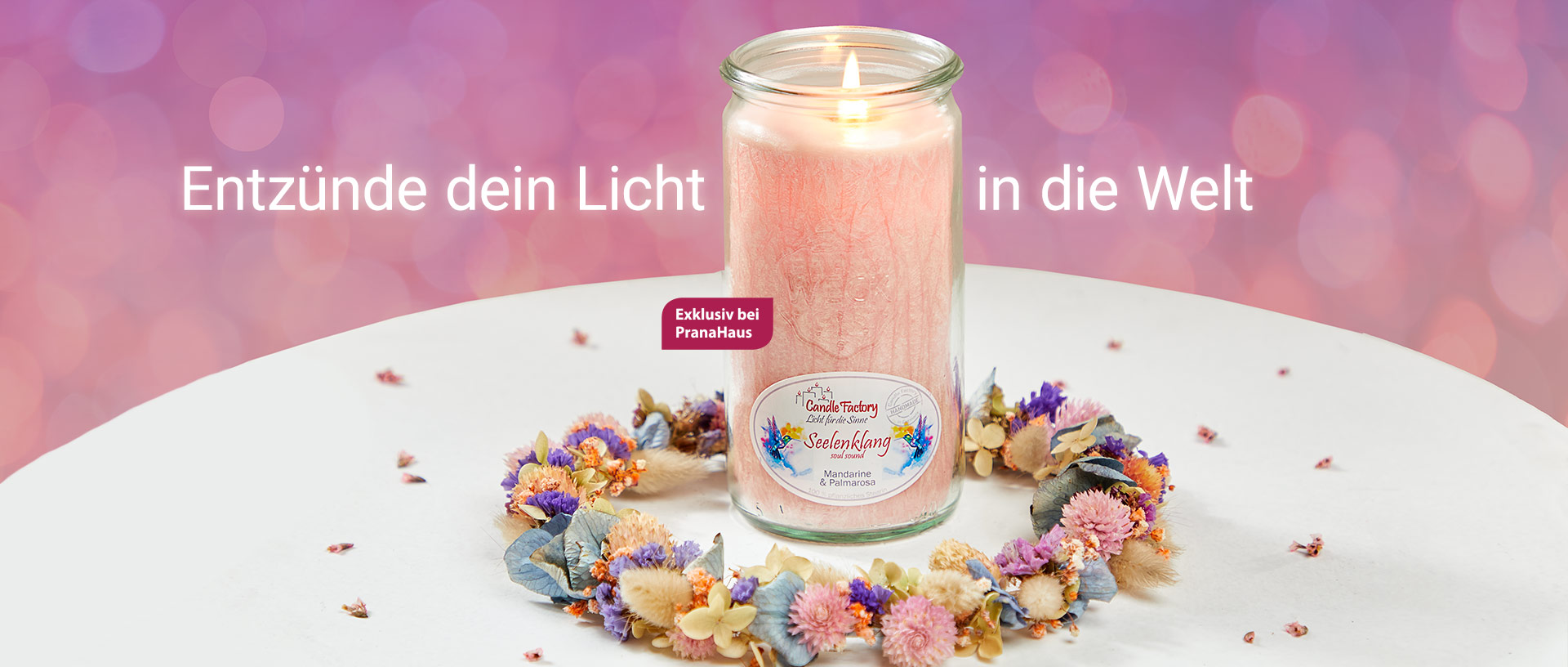 Eine brennende Duftkerze in einem rosa Weckglas steht auf einem Tisch, umgeben von einem bunten Blumenkranz. Daneben stehen die Worte "Entzünde dein Licht in die Welt".