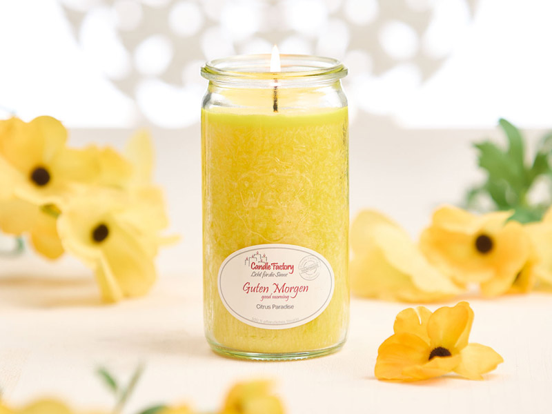 Gelbe Duftkerze im Glas mit Aufschrift „Guten Morgen – Citrus Paradise“, dekoriert mit leuchtenden gelben Blüten, hell und frisch inszeniert.