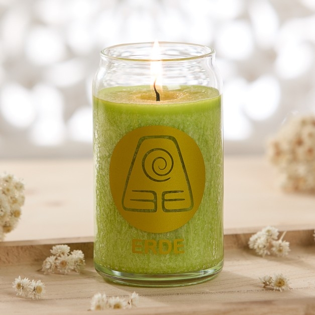 Brennende grüne Kerze im Glas mit der Aufschrift „ERDE“ auf einem hellen Holztisch, umgeben von kleinen Trockenblumen.