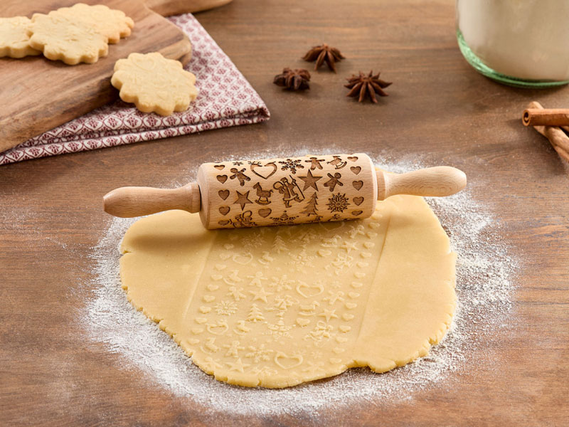 Weihnachtlich gestaltete Teigrolle auf ausgerolltem Plätzchenteig, der mit festlichen Motiven wie Sternen, Herzen und Lebkuchenfiguren geprägt ist.