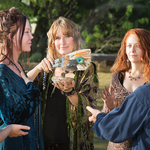 Drei Frauen und ein Mann praktizieren ein Ritual der Wicca-Religion. Sie stehen in einem Kreis, haben die Hände ausgestreckt und halten Schalen und Federn.