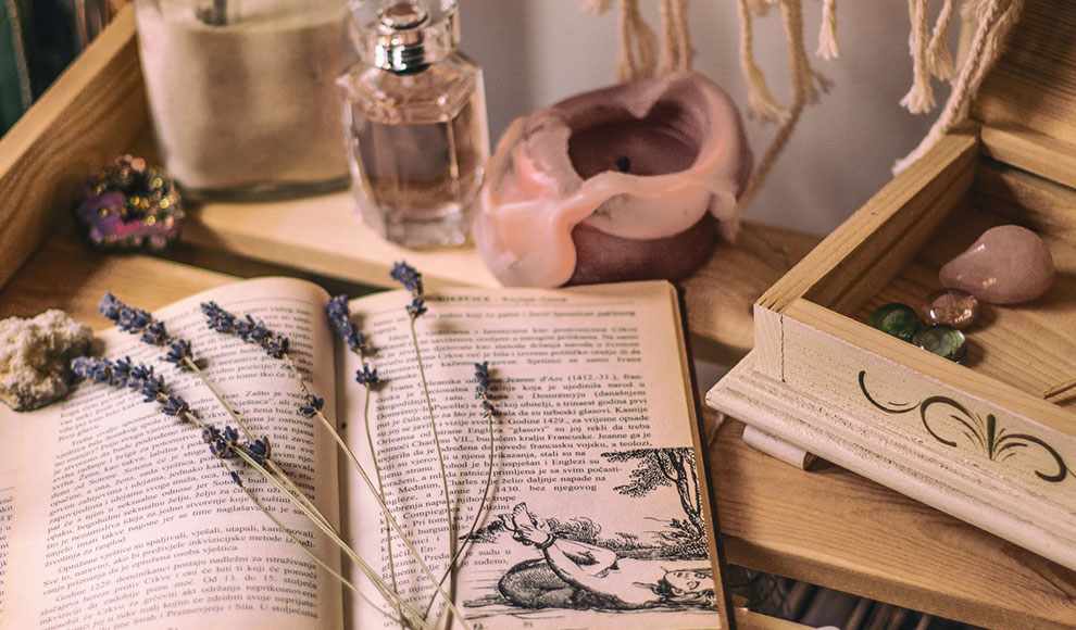 Auf einem Holztisch sind verschiedene Gegenstände angeordnet: Ein Buch, eine Kerze, ein Parfüm-Flakon, mehrere bunte Halbedelsteine, eine Holzschatulle sowie getrockneter Lavendel.