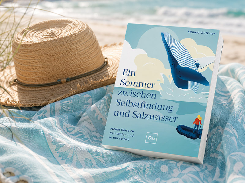 Das Buch "Ein Sommer zwischen Selbstfindung und Salzwasser" liegt auf einerDecke am Strand, mit einem Strohhut im Hintergrund und sommerlicher Meereskulisse.