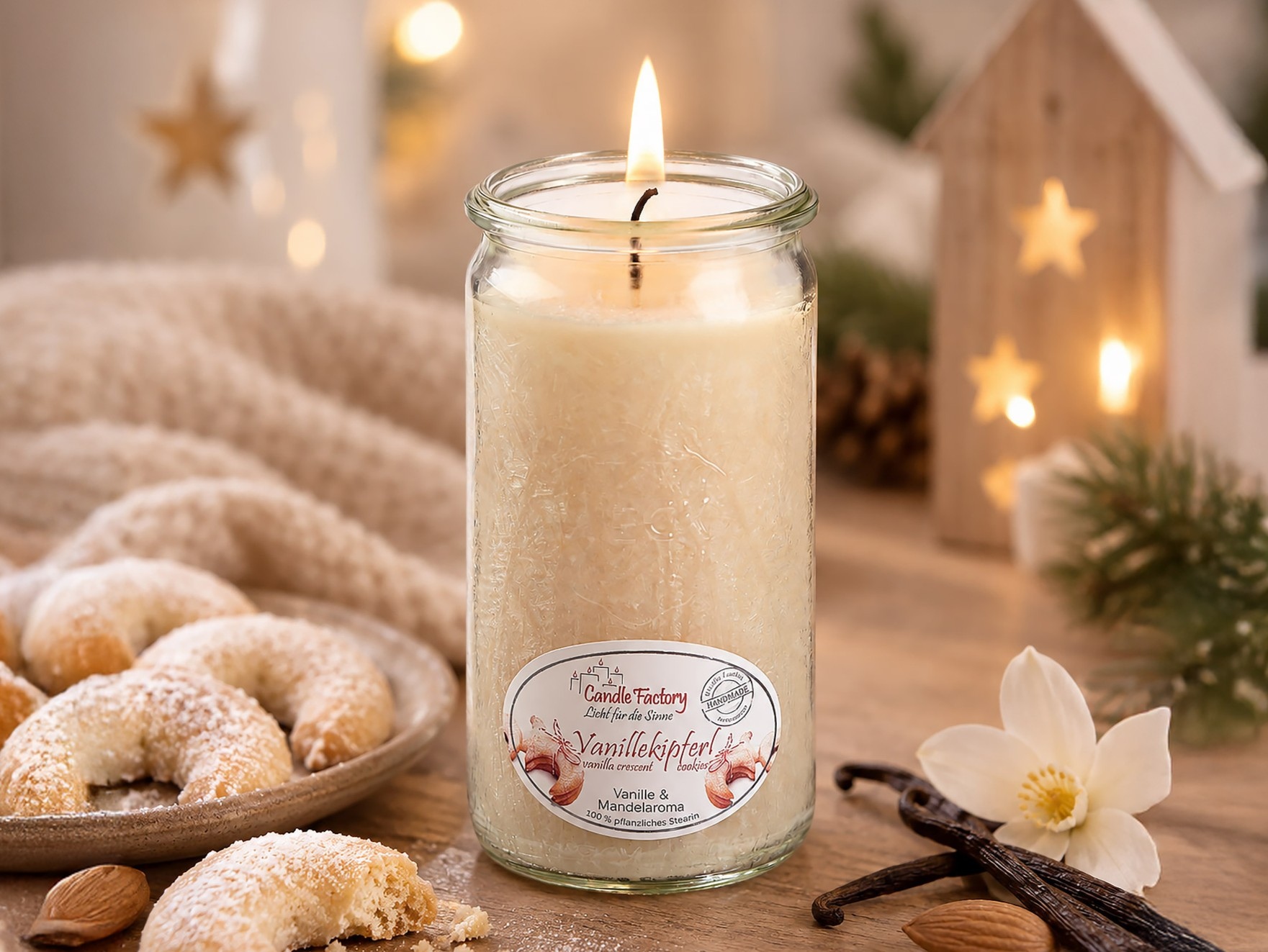 Eine brennende cremefarbene Duftkerze im Glas steht zwischen Vanillekipferln, Vanilleschoten und einer weißen Blüte vor weihnachtlicher Dekoration.