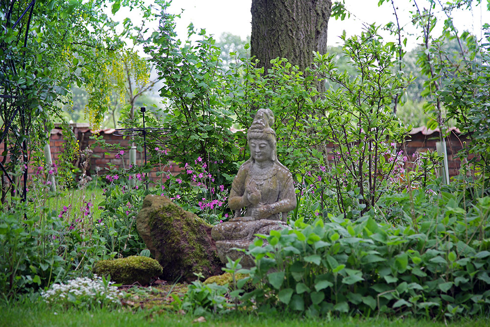 Üppiger Garten mit Grünpflanzen, in dessen Mitte eine steinerne Buddha-Statue sitzt.