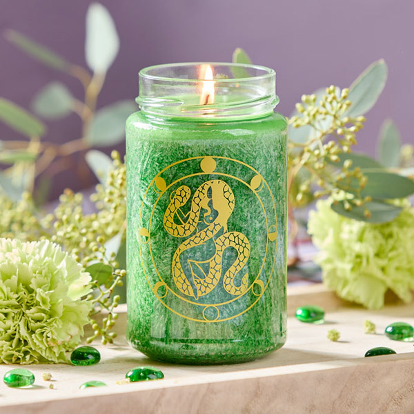 Eine dunkelgrüne, brennende Kerze im Glas mit aufgedrucktem goldenen Mondphasen-Kreis und dem Symbol des Krafttiers Schlange steht auf einem dekorativen Holztablett.