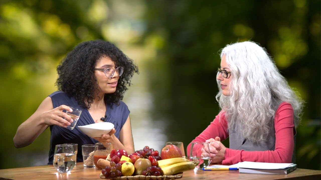 Chantal Sandjon und die Moderatorin sitzen einander zugewandt an einem Tisch, auf dem eine Obstschale steht. Chantal Sandjon hält eine Schüssel und ein Wasserglas in den Händen.