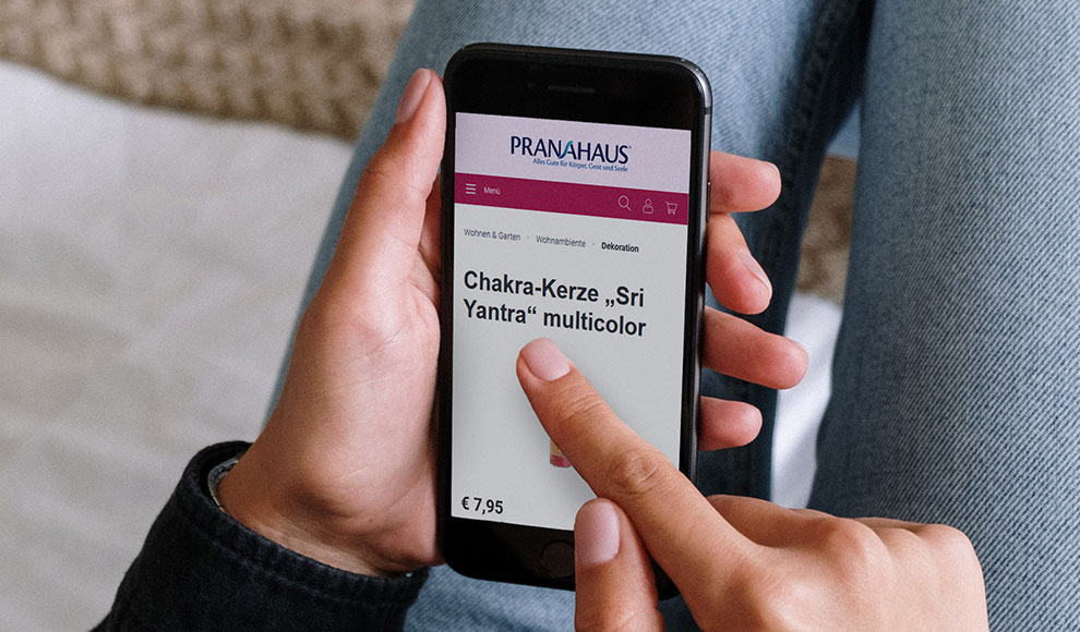 Eine Hand hält ein Smartphone, auf dem der PranaHaus-Shop aufgerufen wurde. Zu sehen ist die Produktdetailseite des Artikels "Chakra-Kerze multicolor".