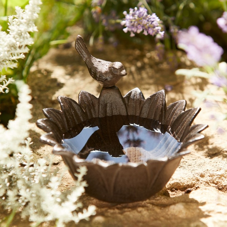 Kleine dunkle Vogeltränke in Blütenform mit einem Vogelaufsatz steht zwischen zarten weißen und lila Gartenblumen.