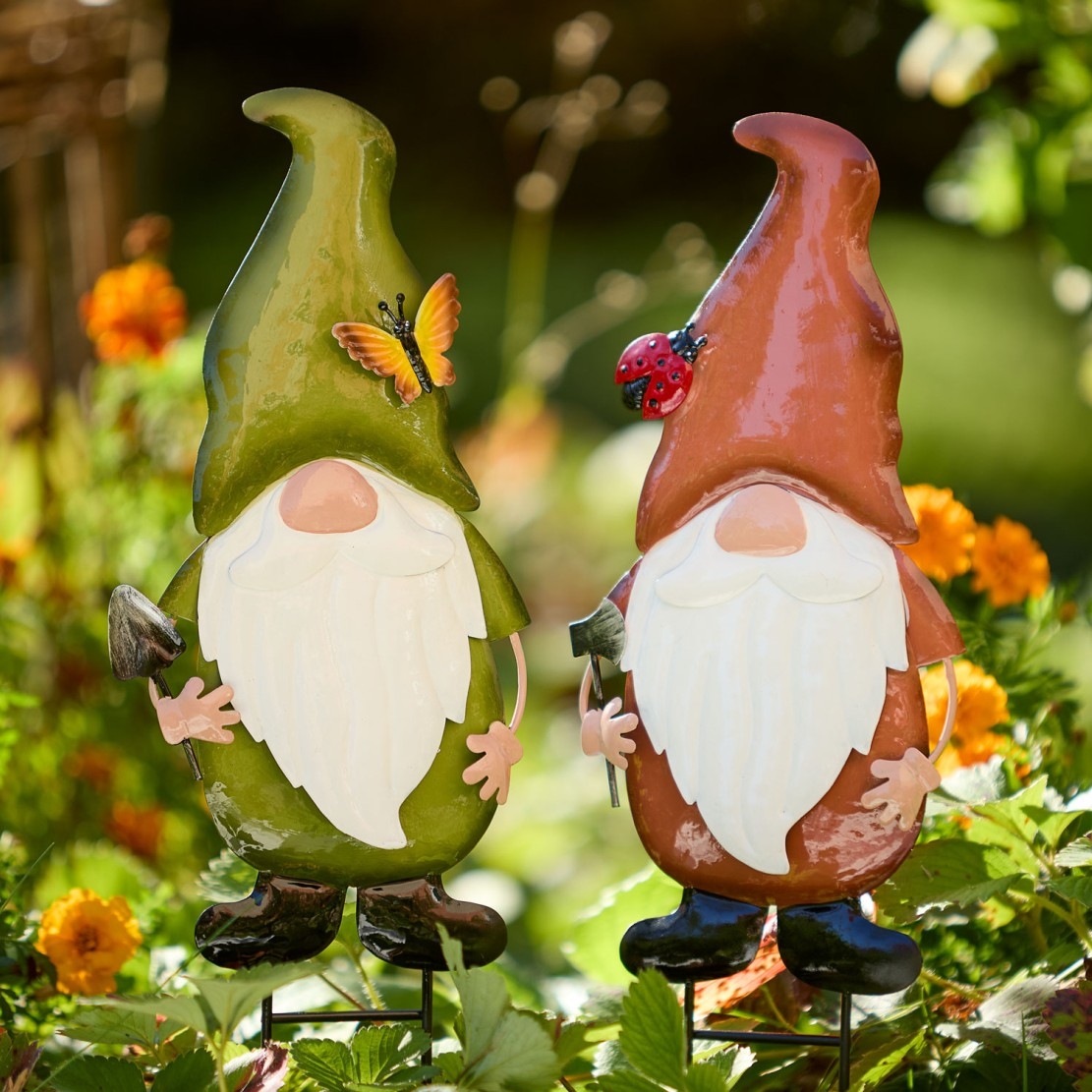 Zwei bunte Gartenwichtel mit langen weißen Bärten stehen zwischen Blumen im Garten und halten kleine Schaufeln.