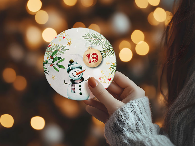 Nahaufnahme einer Hand, die einen runden Adventskalender-Button mit der Zahl 19 und winterlichem Motiv hält.