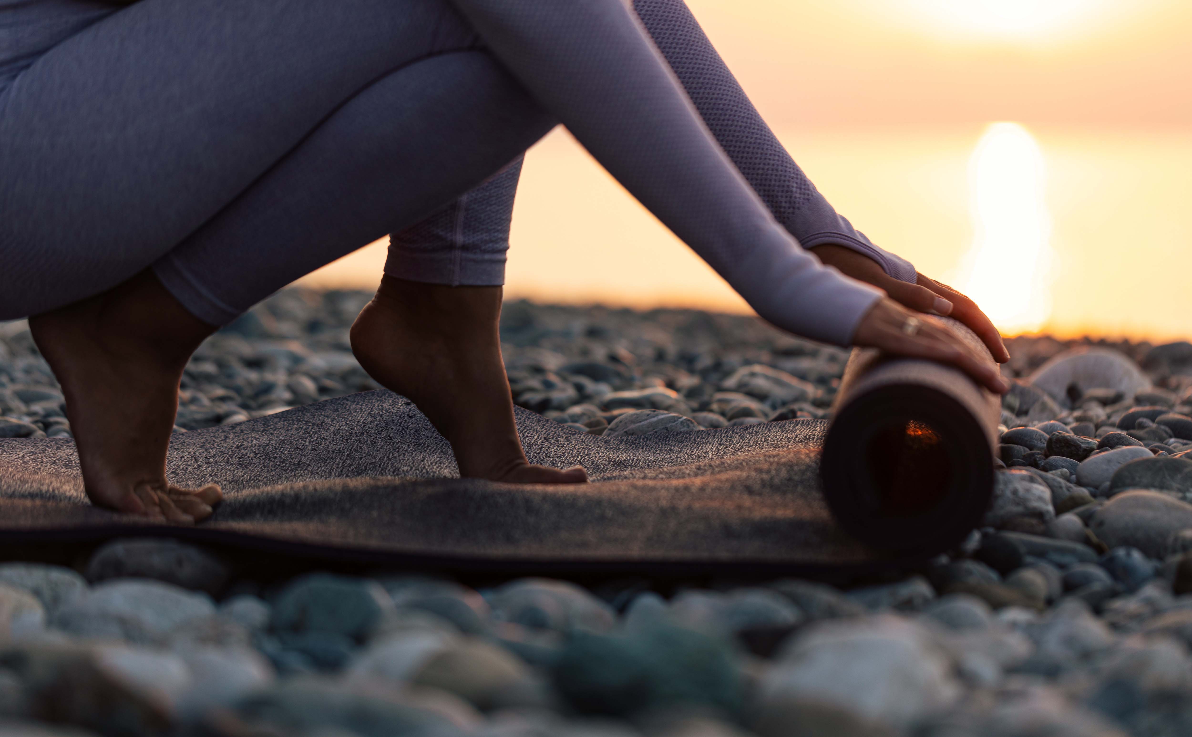 Eine Person in Sportkleidung rollt auf einem Kiesstrand bei Sonnenuntergang eine Yogamatte zusammen.