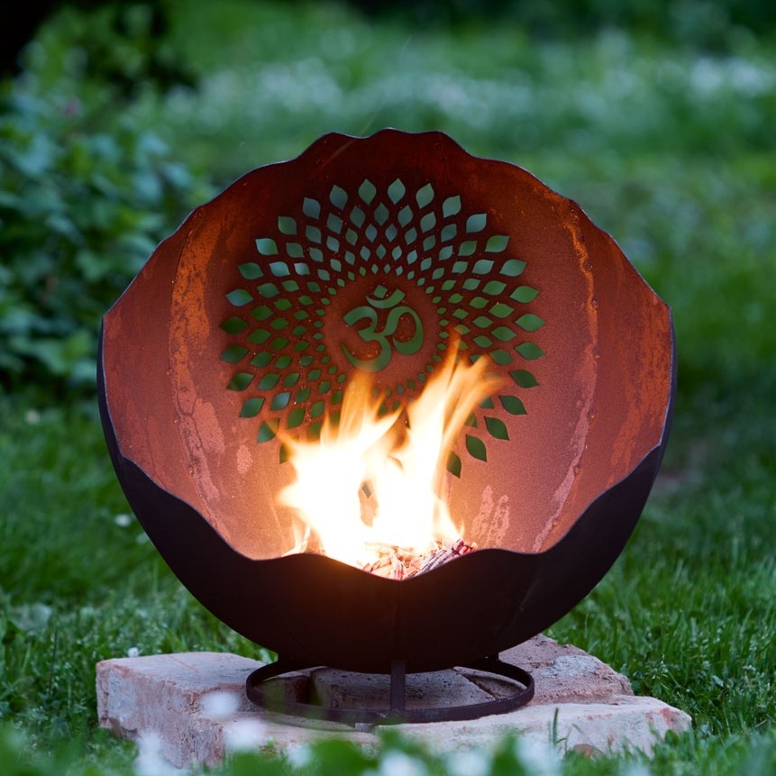 Runde Feuerschale aus rostigem Metall mit dekorativem Muster und Om-Symbol, aus der im Garten Flammen aufsteigen.