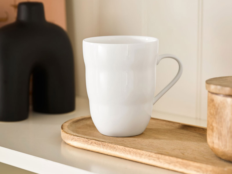 Eine schlichte, formschöne weiße Tasse steht auf einem kleinen ovalen Holztablett in einem Regal. Die Szene vermittelt ein Gefühl von Ruhe – perfekt für eine bewusste Tee- oder Kaffeepause. 