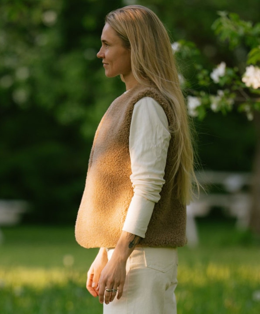 Eine blonde Frau mit langen, offenen Haaren trägt einen beigen Merino-Strickpullunder über einem cremefarbenen Langarmshirt und steht seitlich auf einer grünen Wiese.