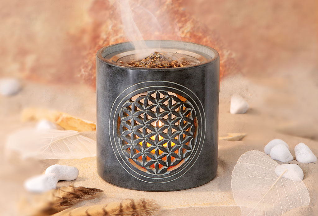 Ein stilvolles Räucherstövchen aus dunklem Stein mit eingravierter Blume des Lebens, durch deren Muster warmes Licht leuchtet. Auf dem Sieb liegt duftendes Räucherwerk, aus dem feiner Rauch aufsteigt.