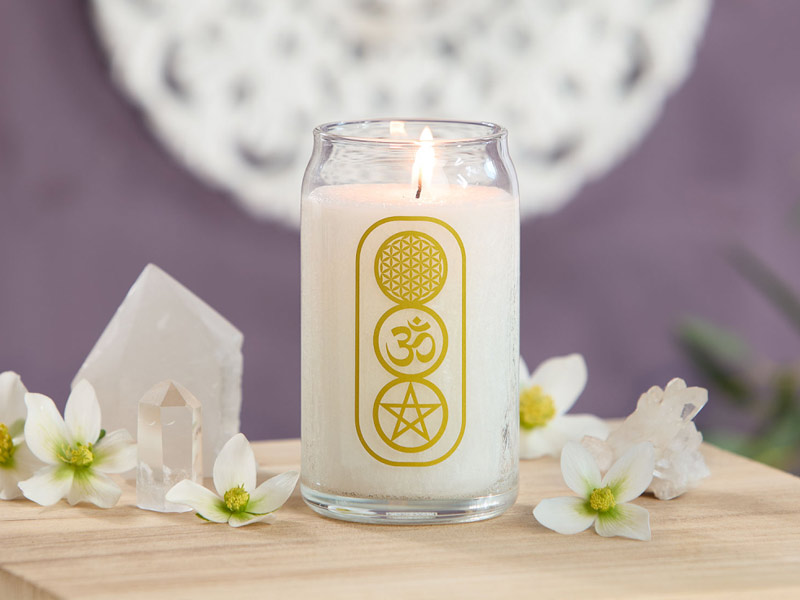 Weiße Duftkerze im Glas mit goldenen spirituellen Symbolen, umgeben von Blüten und Kristallen, in ruhiger, harmonischer Atmosphäre dekoriert.