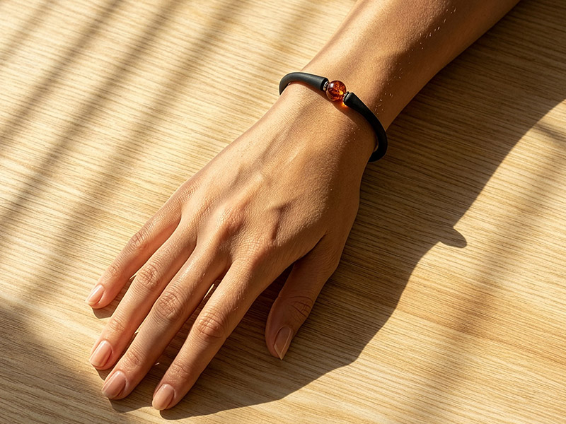 Hand auf hellem Holztisch im warmen Sonnenlicht. Am Handgelenk ein schwarzes Armband mit einer einzelnen Bernstein-Perle, während sanfte Schatten von Fensterstreben über die Szene fallen.