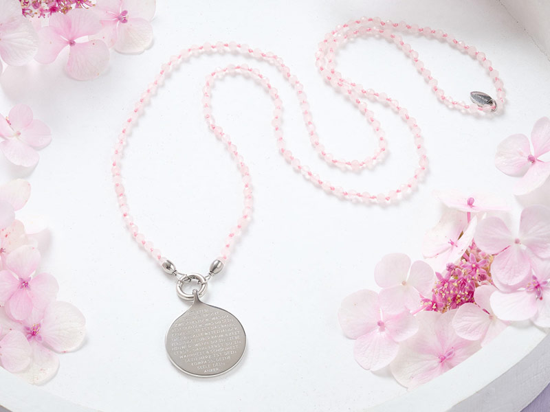 Eine zarte Rosenquarz-Kette mit feinen, facettierten Perlen und einem silbernen, tropfenförmigen Anhänger, der mit einem feinen Mantra-Text graviert ist.