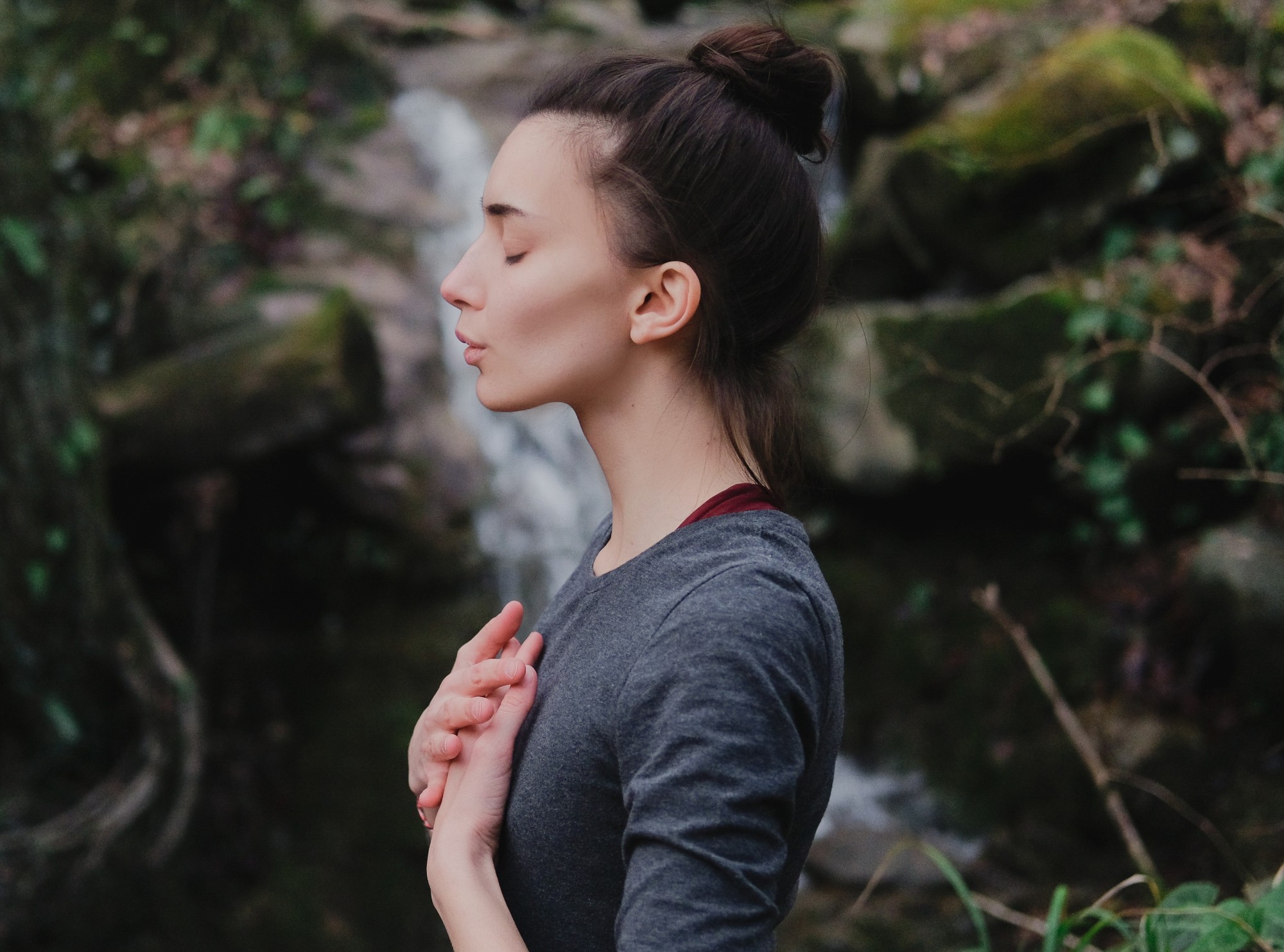 Frau mit geschlossenen Augen hält die Hände vor der Brust und atmet ruhig in einer natürlichen Umgebung am Wasserfall.