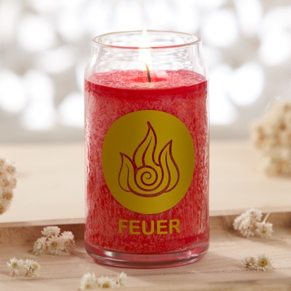 Brennende rote Kerze im Glas mit der Aufschrift „FEUER“ auf einem hellen Holztisch, umgeben von kleinen Trockenblumen.