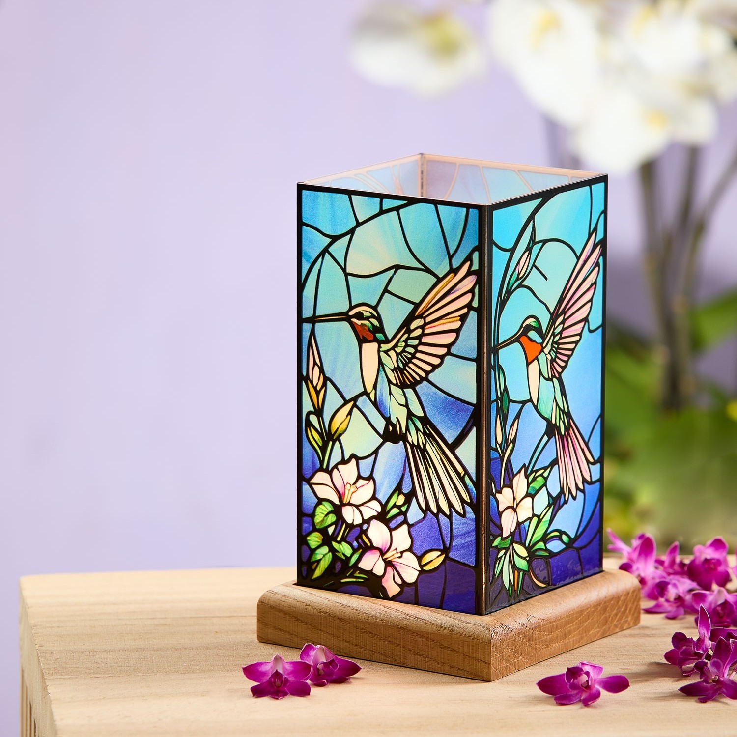 Eine dekorative Tischlampe im Tiffany-Stil mit buntem Glasmotiv von Kolibris und Blumen steht auf einem Holztisch, umgeben von pinken Blüten.