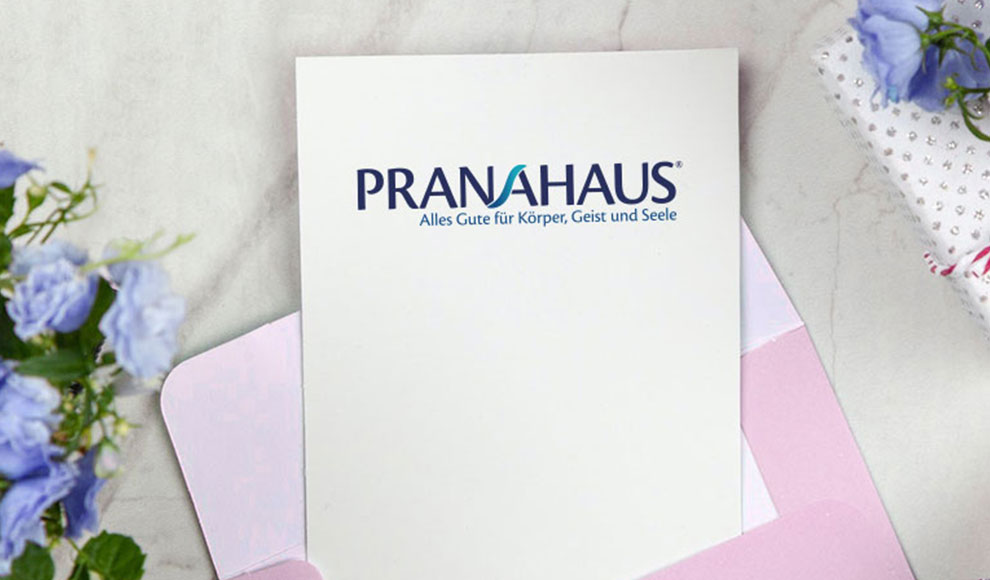 Karte mit PranaHaus-Logo in einem Umschlag, daneben ein Geschenk und ein Blumenstrauß