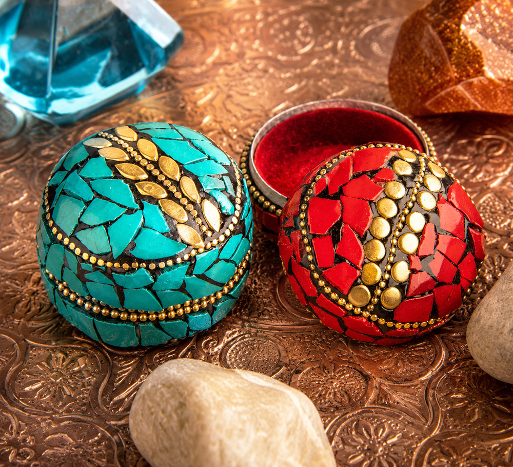 Zwei runde Mosaik-Schmuckdöschen in Türkis und Rot mit goldfarbenen Verzierungen auf einem ornamentierten Kupfertablett. Das rote Döschen ist geöffnet und zeigt sein samtig rotes Innenfutter, umgeben von dekorativen Steinen.