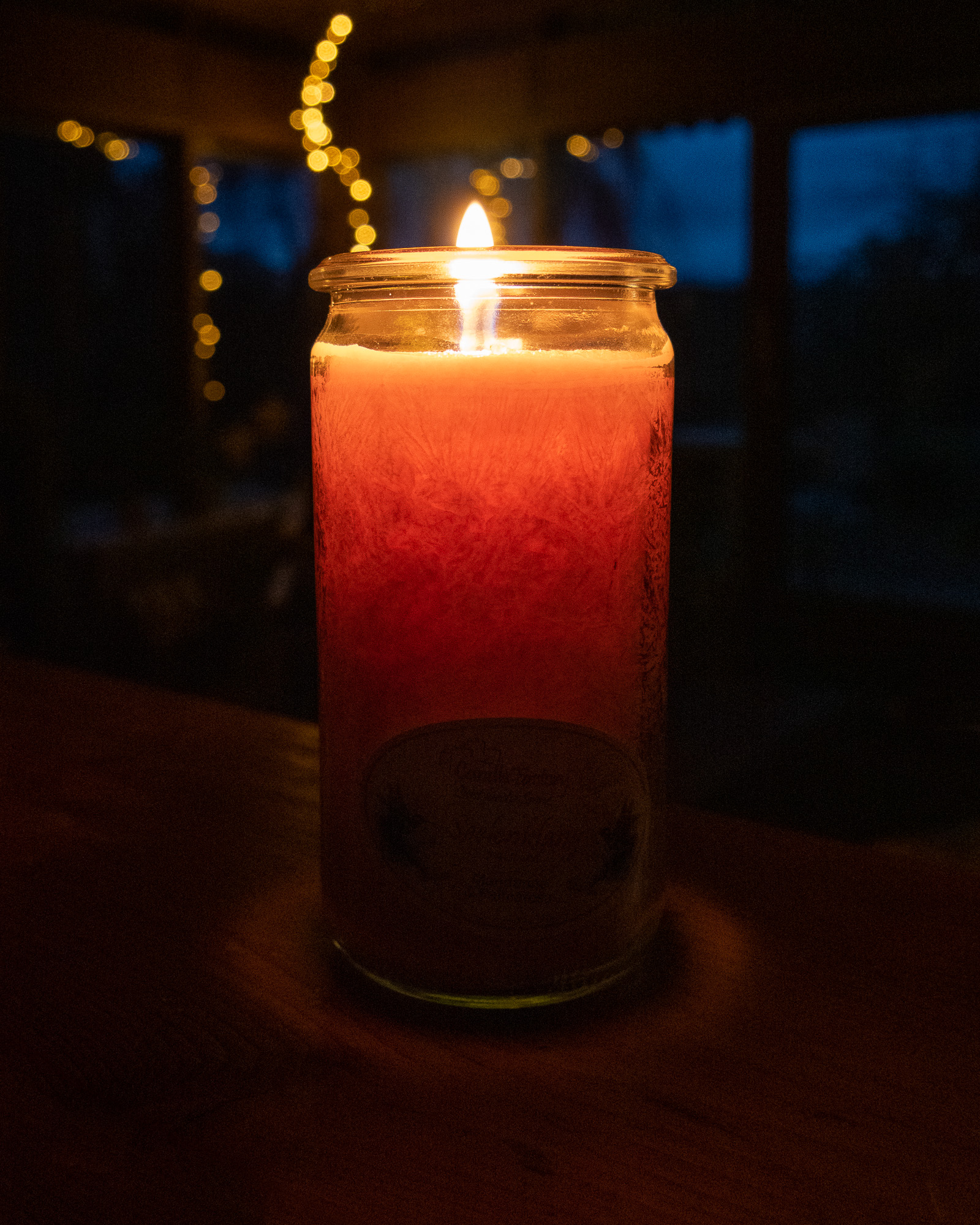 Brennende Kerze auf einem Holztisch, im Hintergrund eine Lichterkette und große Fenster. Es ist blaue Stunde.