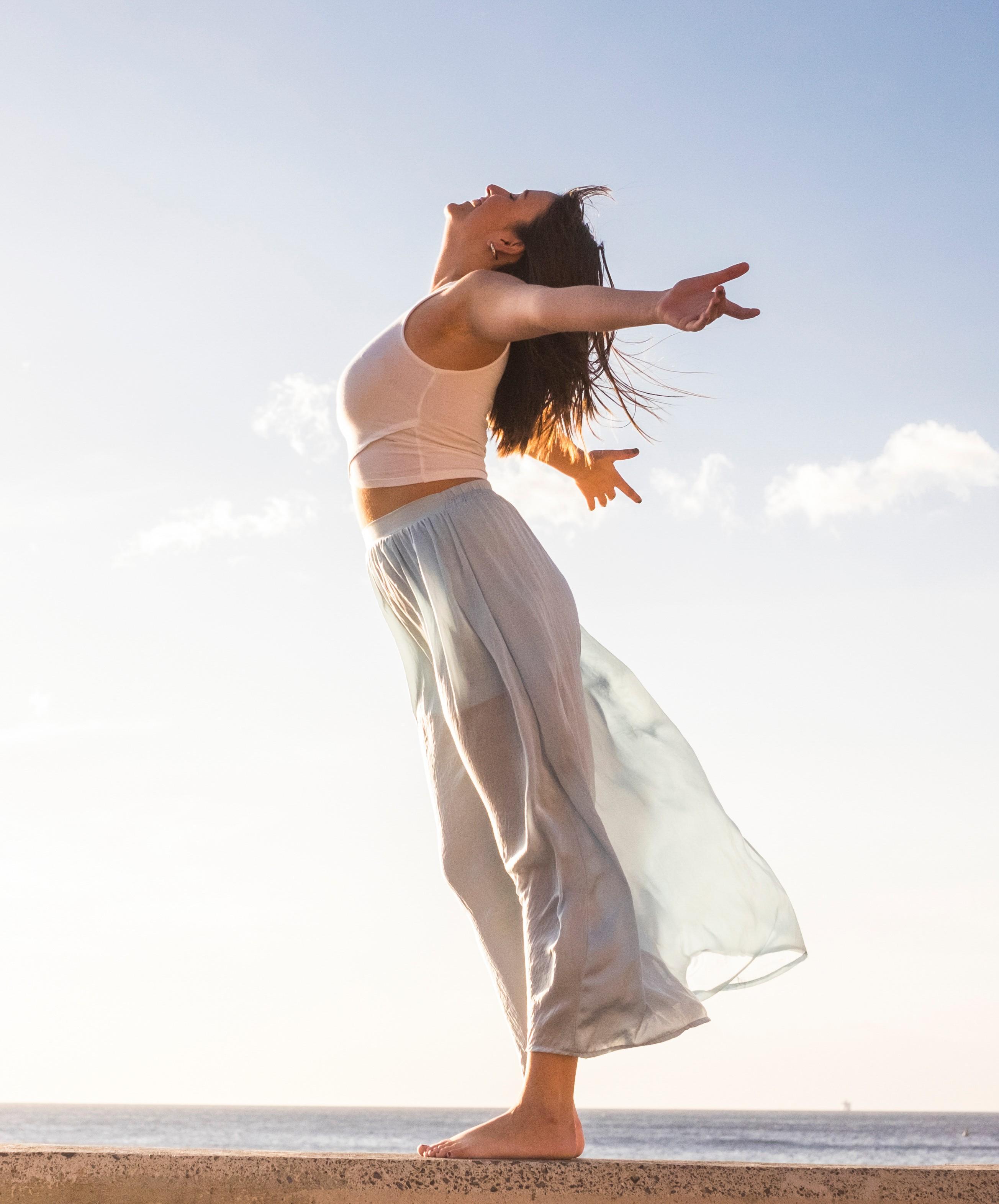 Frau steht mit ausgebreiteten Armen am Meer und wendet das Gesicht entspannt dem Himmel zu.