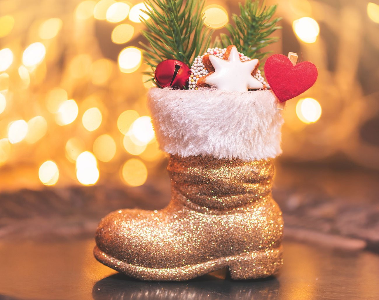 Ein goldglänzender Nikolausstiefel mit weichem, weißem Kunstfellrand steht auf einer dunklen Oberfläche. Er ist festlich gefüllt mit kleinen Tannenzweigen, einer rot glänzenden Glöckchenkugel, einem weiß glasierten Zimtstern und einem roten Filzherz.