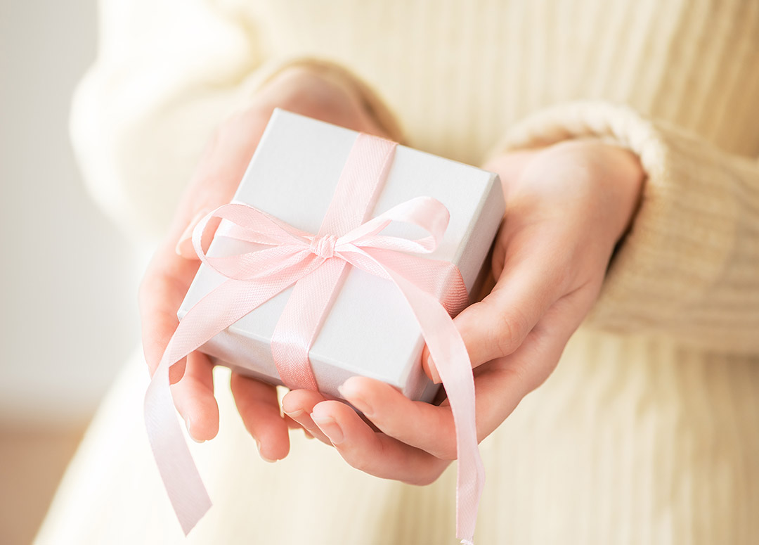 Hände halten ein kleines weißes Geschenk mit rosa Schleife in Richtung Kamera.